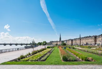 Promenade riveraine à Bordeaux avec espaces verts aménagés, jardins ornementaux et bâtiments historiques en pierre bordant la Garonne sous un ciel bleu dégagé.