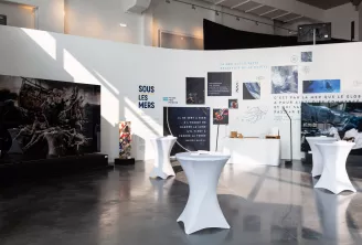 Espace d'exposition contemporain intitulé 'Sous les mers' présentant des tables blanches, des panneaux photographiques sur la conservation marine et des installations textuelles explorant le lien entre l'homme et l'océan.