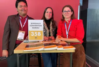 Trois personnes se tenant à un stand « Bordeaux Convention Bureau 35B » lors d'une conférence, portant des badges d'accréditation et présentant des documents promotionnels sur une table en bois devant un fond rose.