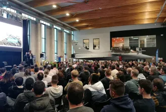 Important groupe de visiteurs réunis dans un auditorium d'Aerocampus Bordeaux, assistant à une présentation sur un avion de chasse militaire exposé sur scène sous un plafond en bois et éclairage industriel.