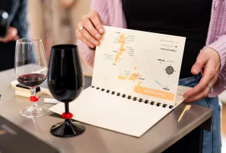 Une femme en cardigan rose tient une carte des régions viticoles devant une table avec des verres de vin rouge et une bouteille, présentant une expérience gastronomique à Bordeaux.