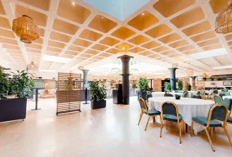 Salle d'événement moderne avec un plafond au design géométrique élégant, tables rondes avec nappes blanches, chaises à cadre doré et assise bleue, séparations en bois, plantes en pot et luminaires suspendus dans un espace lumineux et spacieux.