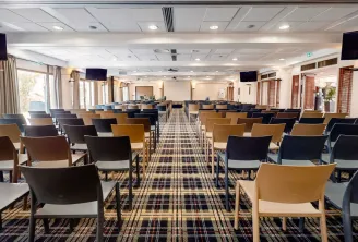 Salle de conférence moderne du Mercure Bordeaux Château Chartrons avec des rangées de chaises en bois et noires disposées en style théâtre, grandes baies vitrées, écrans muraux, et espace de présentation avec écran blanc.