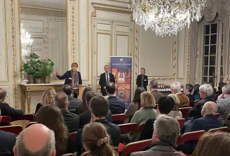 Un événement de présentation formel dans une salle baroque ornée avec lustres en cristal, où trois orateurs se tiennent devant un public assis sur des fauteuils rouges. Une bannière 'Ambassadeurs' est visible, et la décoration met en avant des boiseries c
