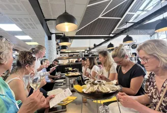 Groupe de personnes dégustant des mets et du vin dans un lieu de restauration moderne avec cuisine ouverte, plafond industriel et luminaires suspendus.