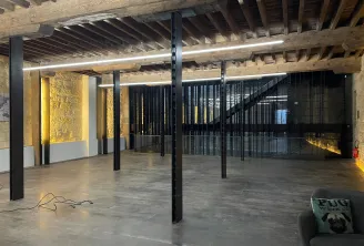 Espace loft industriel moderne avec poutres apparentes en bois, sol en béton, colonnes et garde-corps en acier noir, murs de brique éclairés en jaune, et luminaires contemporains à Bordeaux.