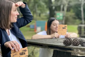 Deux femmes en extérieur dans un parc tenant des cartes illustrées lors d'une activité de team building, avec des pommes de pin sur une table en bois et des arbres en arrière-plan