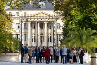 Groupe de professionnels en tenue formelle réuni devant une imposante grille en fer forgé d'une élégante demeure néoclassique bordelaise, avec palmiers et jardins d'ornement.