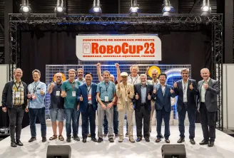 Photo de groupe d'environ 15 personnes posant sur scène à la RoboCup 2023 à Bordeaux, France, les pouces levés en célébration. Une bannière « L'UNIVERSITÉ DE BORDEAUX PRÉSENTE ROBOCUP 23 » est visible en arrière-plan, avec l'éclairage professionnel de la 