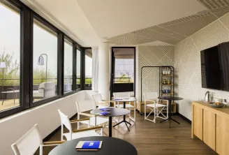 Suite contemporaine de l'hôtel Bordeaux First Name avec un vaste espace de vie, plusieurs coins détente, baies vitrées donnant sur la ville, mobilier design, accents en bois, carrelage à motifs géométriques et chambre adjacente visible.