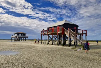 Maisons colorées sur pilotis sur une plage de sable avec des visiteurs explorant le littoral spacieux sous un ciel bleu