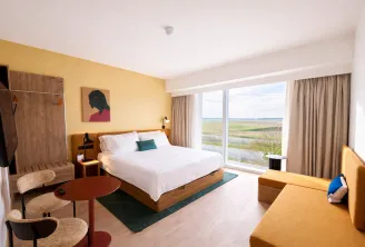 Chambre d'hôtel moderne aux murs jaunes, lit en bois avec draps blancs, grandes fenêtres donnant sur la campagne verte, rideaux beiges, mobilier en bois et décoration contemporaine