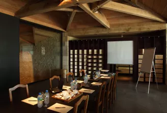 Salle de conférence moderne aux poutres en bois, table longue en bois dressée pour réunion d'affaires avec verres d'eau et sets de place, tableau blanc de présentation, panneaux noirs avec casiers de rangement et éclairage contemporain