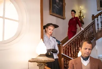 Trois personnes élégamment vêtues dans un escalier majestueux lors d'une enquête immersive au cabaret du Théâtre Molière à Bordeaux. Une femme en veste bleu clair et chapeau noir au premier plan, un homme en veste beige à proximité, et une autre femme en 