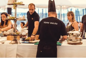 Un chef portant un t-shirt noir 'Team Dulou' se tient à un comptoir de restauration avec des membres de son équipe lors d'un événement à Bordeaux, avec des plats de service et des présentoirs de fruits visibles.