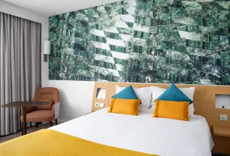 Chambre d'hôtel moderne avec un lit blanc orné de coussins bleus et jaune doré, une tête de lit en bois avec rangements intégrés, un fauteuil en terre cuite, une table de nuit blanche et une grande fresque murale représentant une forêt avec des rayures gé