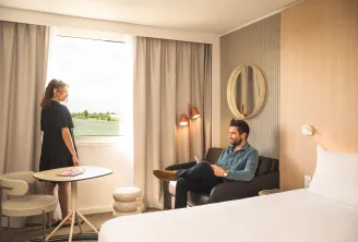 Chambre d'hôtel moderne du Novotel Bordeaux Lac avec deux clients, l'un debout près de la fenêtre donnant sur le lac et l'autre assis sur le canapé, avec mobilier contemporain et éclairage chaleureux