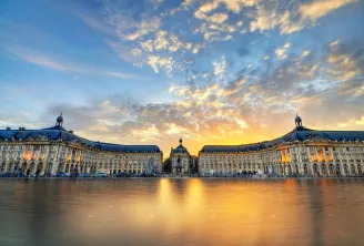 Place de la Bourse à Bordeaux au coucher de soleil, avec ses deux bâtiments symétriques du XVIIIe siècle aux toits mansardés bleus se reflétant dans le miroir d'eau, ciel doré dramatique