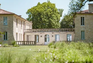 Château en pierre de style classique avec pelouse tondue au premier plan, arbres centenaires et ciel dégagé dans la région viticole de Bordeaux