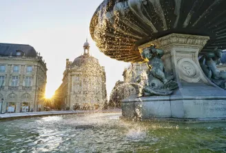 Fontaine dorée avec jets d'eau sur la place historique de Bordeaux au coucher de soleil, avec des bâtiments en pierre classiques et une église à dôme en arrière-plan