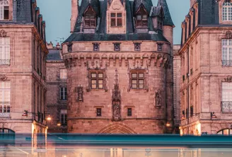 Bâtiment historique en grès rouge avec tours ornementées et toits mansardés à Bordeaux, illuminé au crépuscule avec le trafic flou en avant-plan