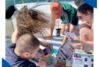 Un groupe de participants à bord d'un voilier consulte des documents et prend des notes lors d'une activité éducative marine dans le Bassin d'Arcachon, avec la mer et l'équipement du bateau en arrière-plan.
