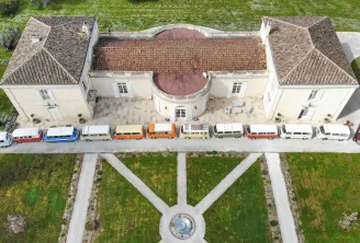Vue aérienne d'une propriété classique française avec cour centrale présentant une file de camping-cars vintage colorés garés devant le bâtiment principal, entourés de jardins aménagés et de chemins géométriques