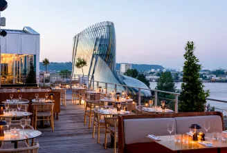 Terrasse élégante de restaurant en toiture au crépuscule surplombant la Garonne à Bordeaux, avec l'architecture distinctive de la Cité du Vin visible en arrière-plan, tables dressées pour le fine dining avec verres et bougies.