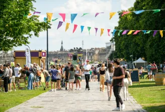 Scène de festival en plein air à Bordeaux avec des guirlandes colorées, des foules de visiteurs circulant sur une allée pavée, des petits stands en bois à gauche, des espaces verts et des monuments historiques de la ville visibles en arrière-plan sous un 