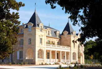 Un château de prestige aux façades en pierre de taille couleur crème, doté de deux tourelles coniques aux toits d'ardoise sombre, de fenêtres cintrées, de balustrades ornementales et d'un portique à colonnes, entouré de verdure sous un ciel bleu dégagé.