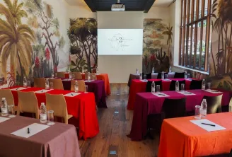 Salle de conférence contemporaine de l'Hôtel La Zoologie à Bordeaux avec des nappes multicolores rouge, orange et bordeaux, des chaises en bois, des bouteilles d'eau, des fresques murales botaniques tropicales, un écran de projection et de grandes fenêtre