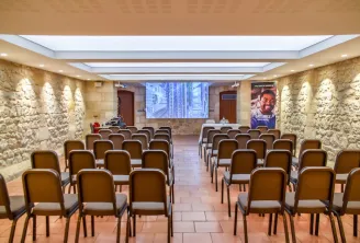 Salle de conférence moderne avec chaises rembourrées marron alignées en rangées, murs en pierre, plafond avec puits de lumière et éclairage LED, écran de projection affichant une vue urbaine au Château Grattequina à Bordeaux.
