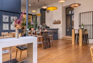 Espace bar et salon contemporain d'un hôtel boutique avec sol en bois, luminaires suspendus design, comptoir blanc avec tabourets, bar arrière sombre garni de bouteilles, tables de repas en bois avec chaises, arrangements floraux frais et grandes baies vi