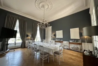 Salle à manger élégante du Château Gassies à Bordeaux, avec murs gris anthracite, rosace plafond blanche ornementée, lustre en cristal, table de repas blanche, chaises grises capitonnées, grandes fenêtres aux rideaux gris, parquet point de Hongrie et mobi
