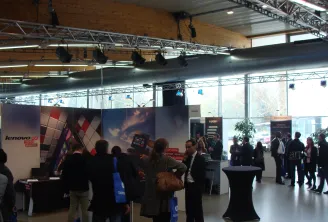 Salon professionnel intérieur avec stands d'exposition Lenovo, visiteurs en tenue professionnelle, et infrastructure moderne avec charpentes métalliques et panneaux d'affichage grand format.