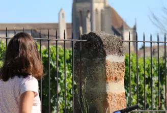 Une jeune femme à vélo s'arrête devant une clôture en fer donnant sur des vignobles, avec le clocher d'une église historique en arrière-plan à Saint-Emilion.