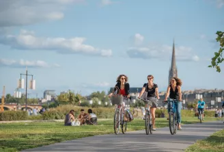 Trois femmes à vélo sur une piste cyclable ombragée dans un parc verdoyant avec la cathédrale de Bordeaux et ses toits en arrière-plan, tandis que d'autres visiteurs profitent de l'espace.