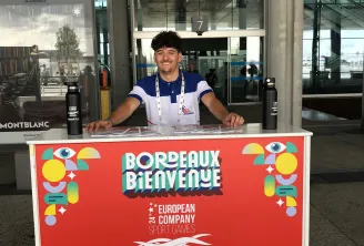 Un jeune homme en uniforme de bénévole bleu et blanc se tient à un stand d'accueil affichant une bannière rouge souhaitant la bienvenue aux visiteurs des Jeux du Sport d'Entreprise Européens à Bordeaux, du 14 au 18 juin, au Hangar 14 et dans plus de 30 si