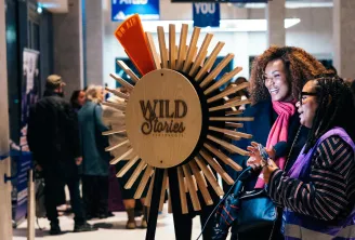 Deux visiteurs observent une grande installation interactive en forme de soleil doré portant le label 'Wild Stories' dans un espace moderne à Bordeaux, avec des écrans numériques visibles en arrière-plan.