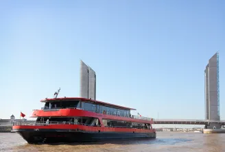 Bateau de transport de passagers rouge et bleu naviguant sur la Garonne à Bordeaux, avec des tours jumelles modernes et un pont visible en arrière-plan sous un ciel bleu dégagé.