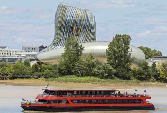 Un bateau de croisière rouge et noir naviguant sur la Garonne à Bordeaux, avec l'architecture moderne et courbe de la Cité du Vin visible en arrière-plan sous un ciel bleu parsemé de nuages blancs.