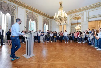 Un homme parlant à un pupitre s'adresse à un grand groupe de participants réunis dans une salle de palais ornée, décorée de dorures, lustres en cristal et parquet, lors d'une journée éducationnelle à Bordeaux.