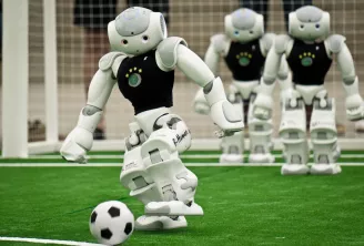 Robot humanoïde en maillot noir frappant un ballon de football sur un terrain vert, trois robots identiques visibles en arrière-plan, illustrant l'excellence en robotique.