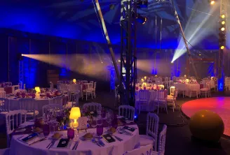 Salle d'événement élégante avec tables rondes dressées pour un dîner, décorée d'éclairages chauds et bleus ambiants, sous une structure de chapiteau de cirque avec éclairages professionnels et piste de danse rouge