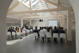 Une présentation professionnelle dans une salle de conférence moderne de style loft avec poutres en bois apparentes et verrière. Environ 30 participants professionnels assis sur des chaises blanches face à un présentateur près d'un écran de projection.