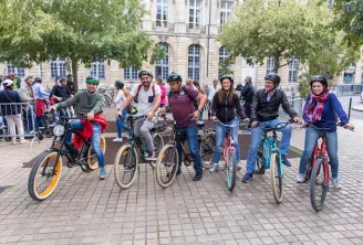 Un groupe de cyclistes équipés de casques pose ensemble sur une place pavée devant un bâtiment en pierre classique, lors d'une balade à vélo urbaine à Bordeaux.