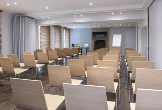Salle de conférence contemporaine de l'hôtel Seeko'o à Bordeaux avec des rangées de chaises en bois clair munies de coussinets beige, équipées de surfaces de prise de notes, face à une zone de présentation avec écran affichant le logo Seeko'o, éclairage l