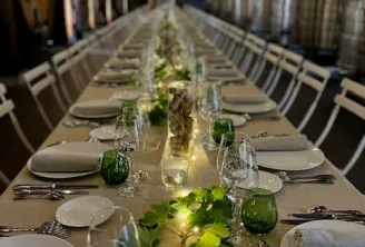 Table de banquet allongée dressée pour un dîner raffiné avec assiettes blanches, verres et un décor floral vert au centre, éclairée par des lumières suspendues dans une salle moderne.