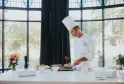 Chef professionnel en uniforme blanc et toque préparant des canapés et amuse-bouches raffinés à un comptoir moderne avec baies vitrées donnant sur un jardin, avec des fleurs fraîches et des ingrédients en vitrine.