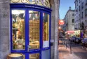 La devanture bleue élégante de la boutique Cousin & Compagnie à Bordeaux, avec une ferronnerie ornementale au-dessus des vitrines et un tonneau en bois à l'extérieur, située dans une rue historique bordée de bâtiments traditionnels.